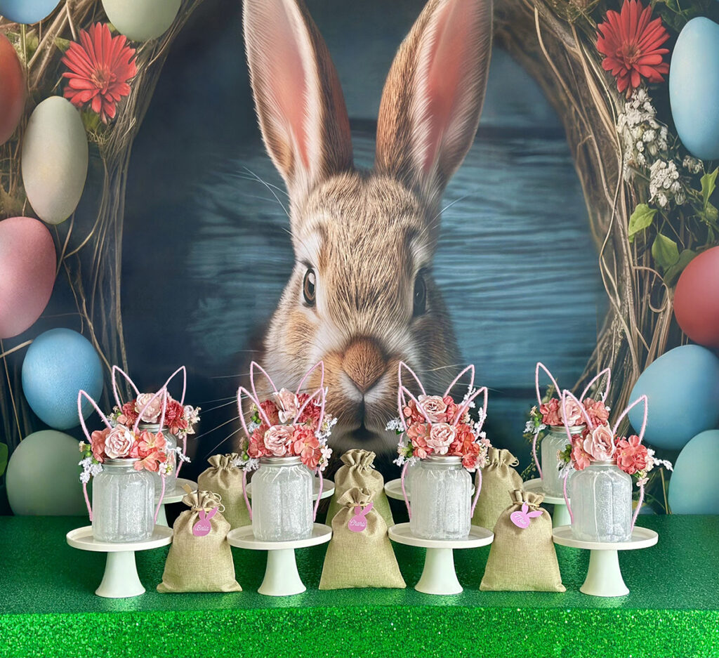 Easter Fairy Jar Lantern Party table display with tissue-covered mason jars topped with pink floral bunny ear headbands, hessian favour bags with pink bunny tags, and a large backdrop of a realistic bunny surrounded by colourful Easter eggs and flowers on a green sequin tablecloth.