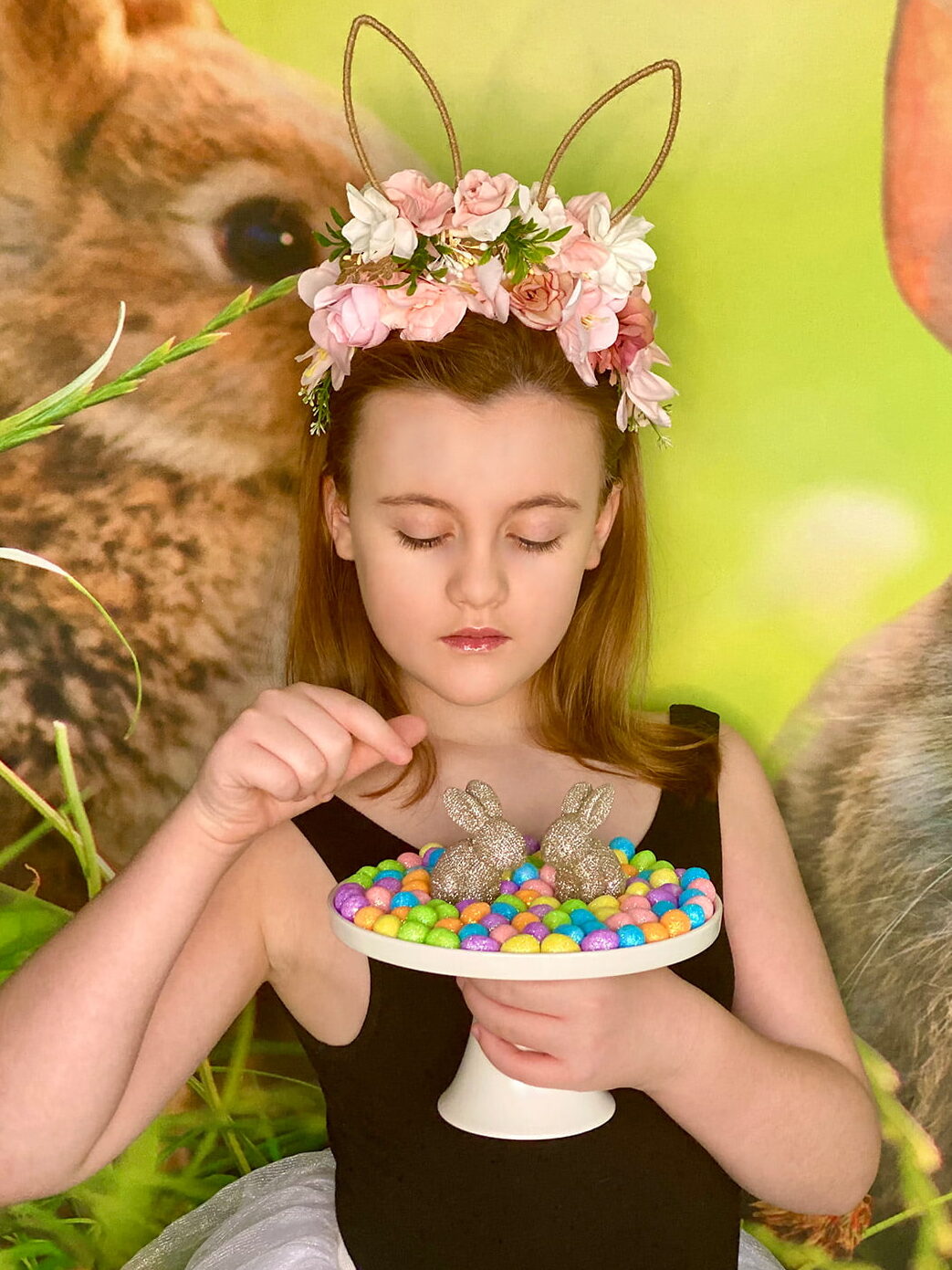 Young girl wearing a bunny ear headband, holding a white cake stand with gold bunnies and pastel eggs, looking down sweetly as if about to pick up an egg.