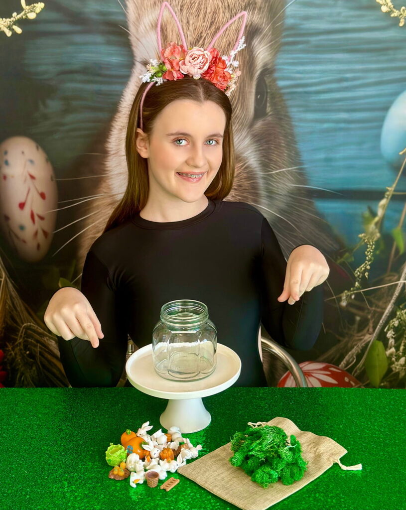 Girl pointing with both hands at an empty mason jar on a cake stand in front of her, with Easter terrarium decorations arranged on a hessian bag atop a green sequin tablecloth, ready to start crafting.