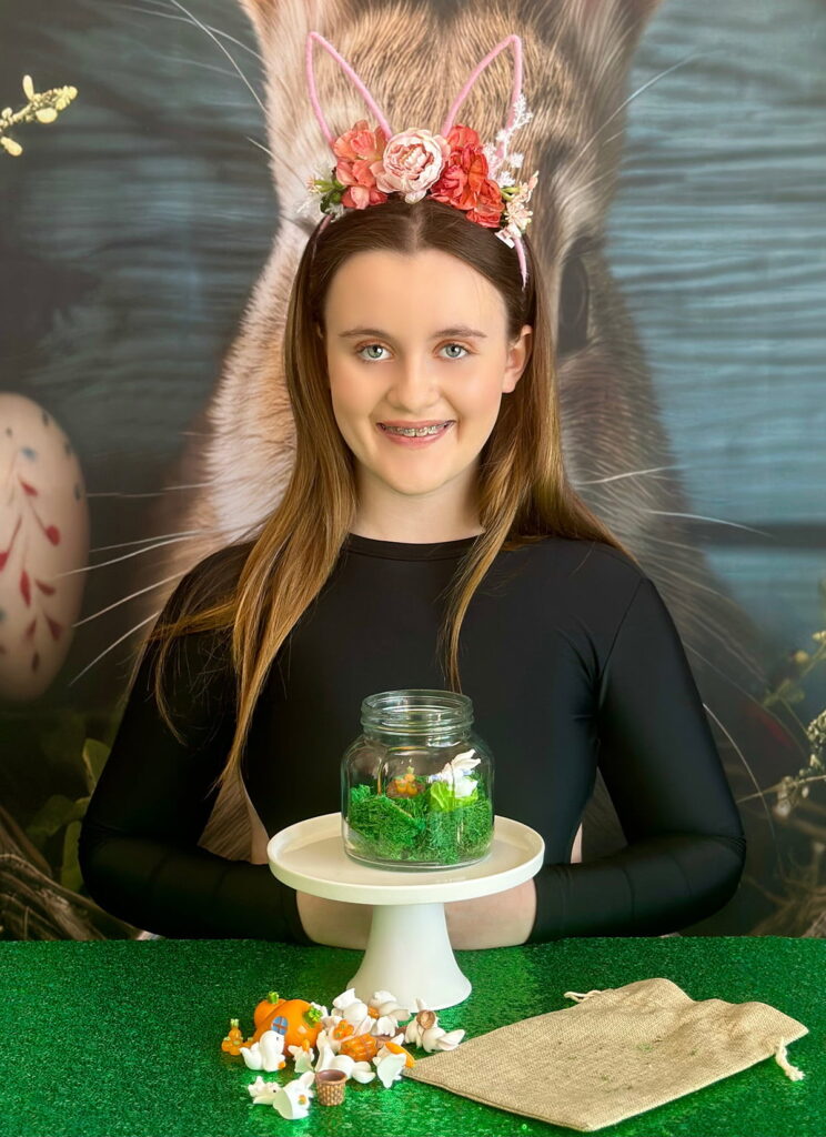 miling party girl decorating her Easter terrarium, with a moss-filled mason jar on a white cake stand and bunny and carrot miniatures beside it on a hessian bag.