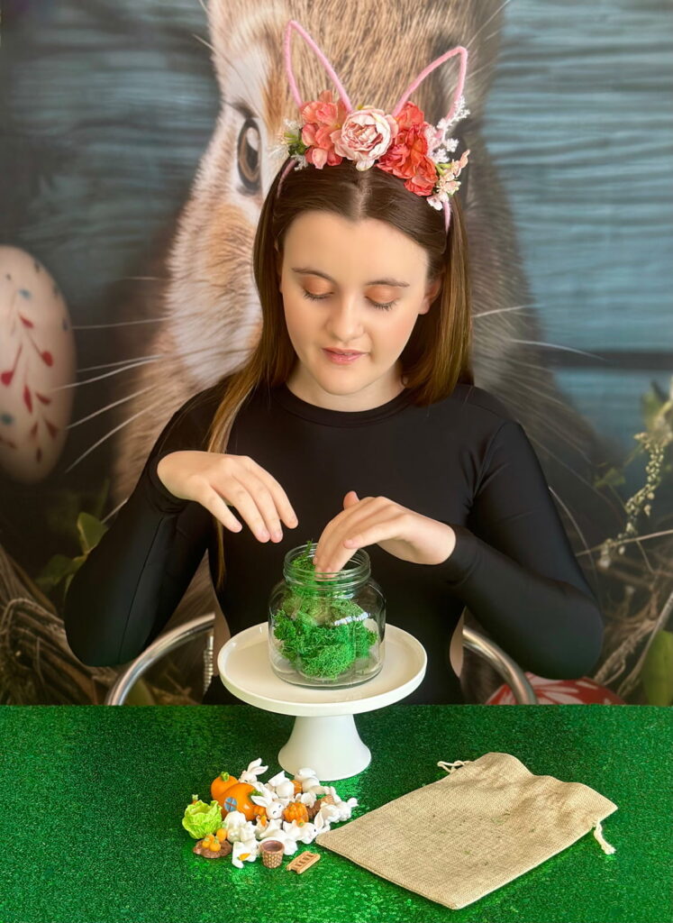 Young girl at the Easter terrarium party table placing faux green moss inside a clear mason jar on a white cake stand, with a hessian bag and party decorations in front of her.