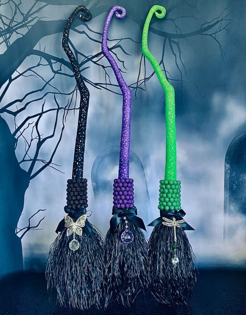 Three decorated witches’ brooms in black, green, and purple on a table with a spooky graveyard and tree backdrop, part of a kids’ witch-themed party activity.