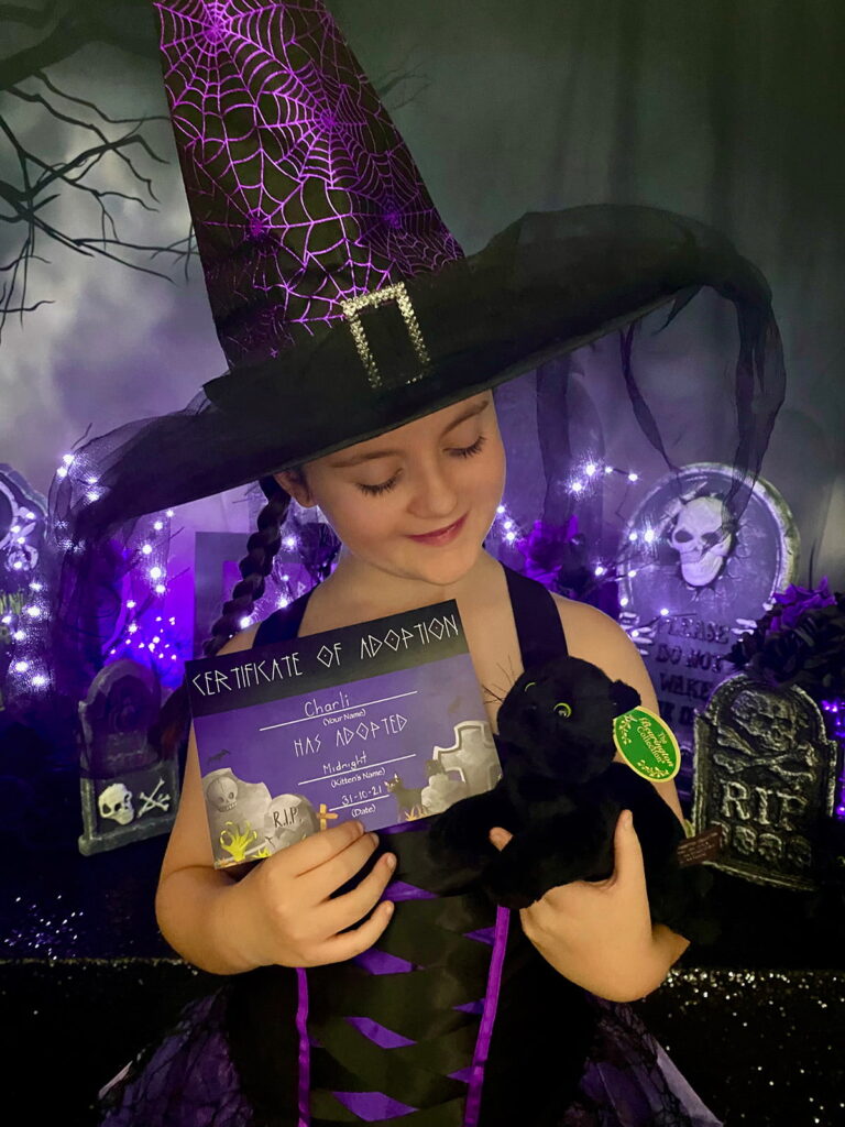 Young girl in a purple and black witch hat and dress standing in front of a kitten adoption party table, holding an adoption certificate in one hand and a plush black kitten in the other, smiling while looking down at her adopted kitten.