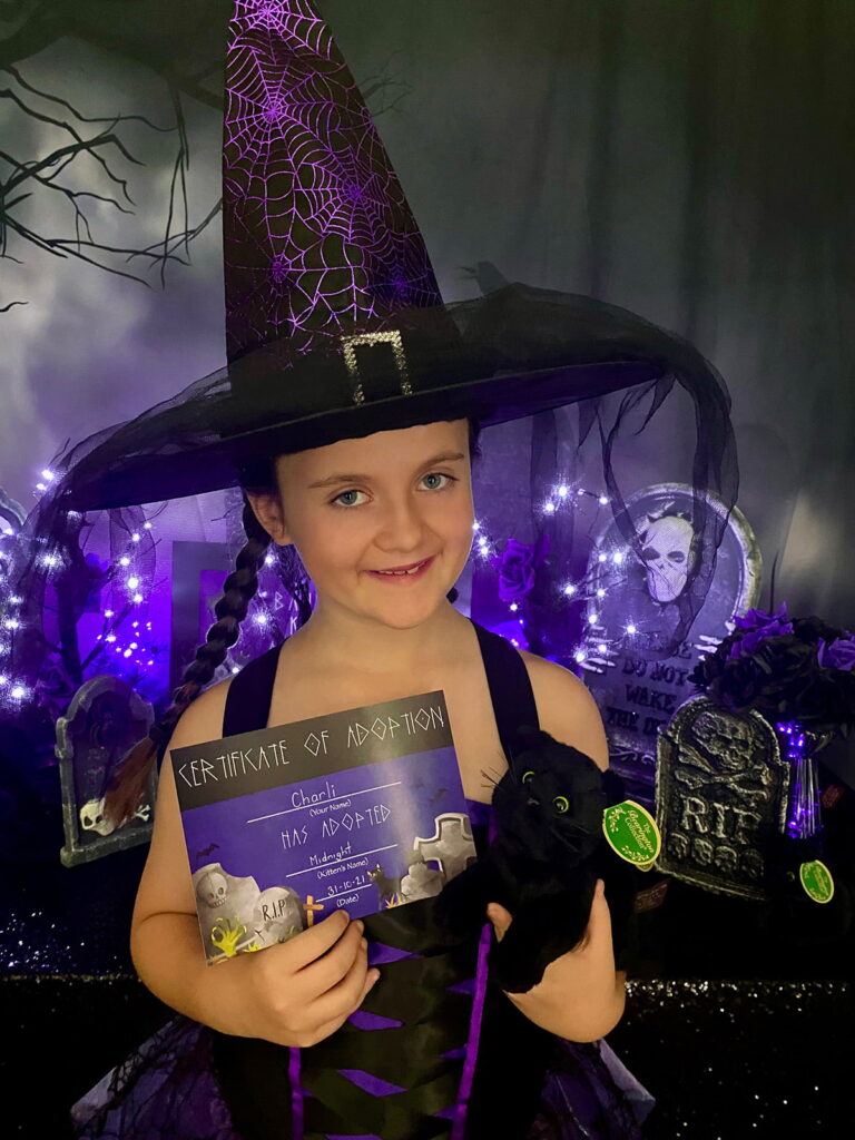 Young girl in a large purple witch hat and dress standing in front of a kitten adoption party table, smiling at the camera while holding an adoption certificate and a plush black kitten after adopting it at a kids’ witch-themed party.