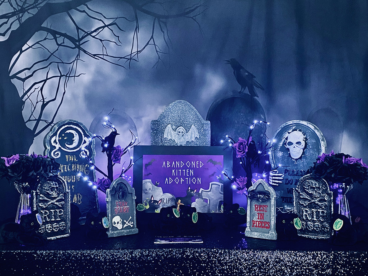 Spooky-cute kitten adoption party table for kids with a graveyard backdrop, black sequin tablecloth, and plush black kittens nestled among tombstones and flowers under glowing purple lights for a witch-themed activity.