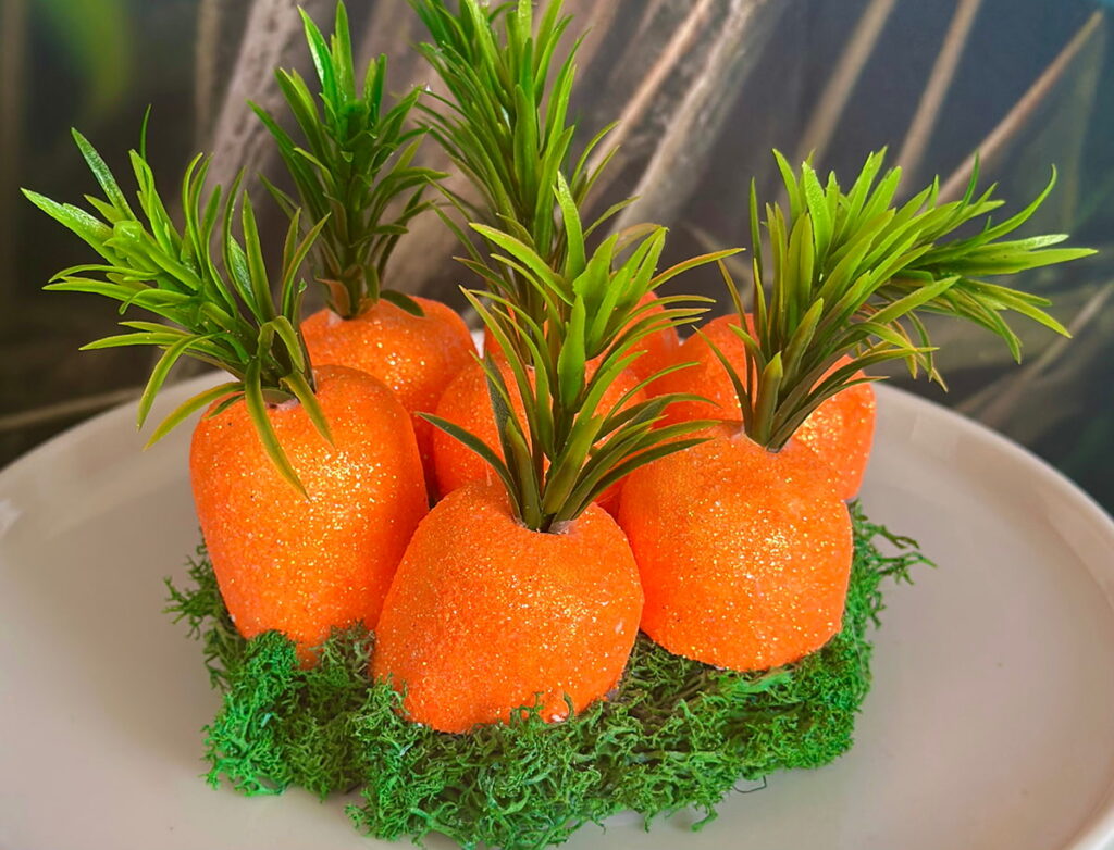 Six bright orange foam carrot tops glued on a moss-covered terrarium lid, displayed neatly on a white cake stand against a soft background.