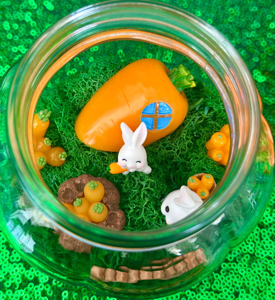 Overhead view of a decorated Easter terrarium with a carrot-shaped house, bunny figurines, carrot decorations, a small brown fence, and green moss.