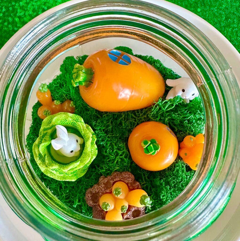 Top-down view inside an Easter terrarium showing bright green moss, a miniature carrot house, small bunny figurines, carrot decorations, and a tiny bunny nestled in lettuce leaves.