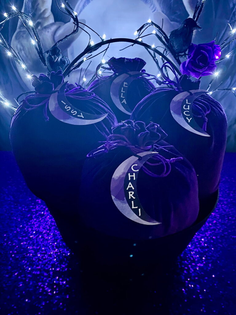 Purple velvet bags filled with crystal ball decorating supplies, arranged in a large black cauldron and labelled with moon-shaped name tags for a witch-themed kids party.