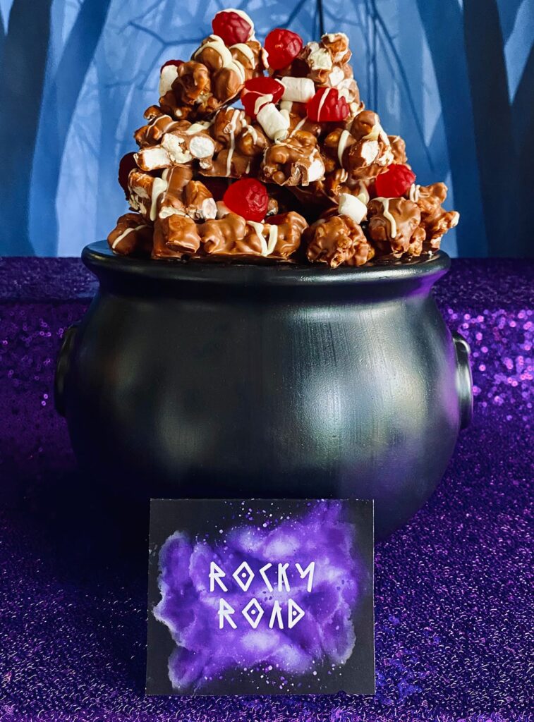 Black cauldron filled with rocky road popcorn piled high with chocolate-coated popcorn, gummy raspberries, and marshmallows, served as a spooky treat for a witch-themed kids’ party.