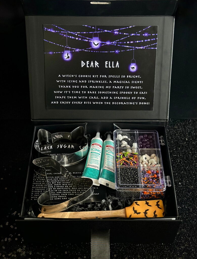 Witch cookie kit party favour in a black gift box with black shredded paper, containing witch-themed cookie cutters, icing pens, themed sprinkles, an orange mini spatula with a bat design, and a black sugar cookie recipe card.