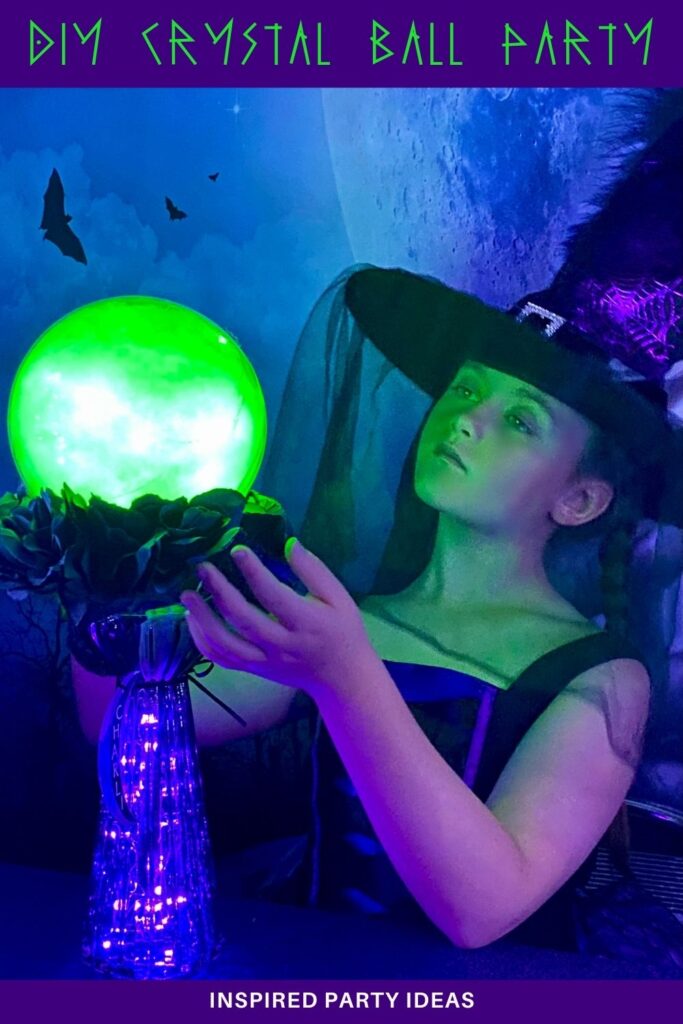 Young girl wearing a witch hat with a veil over her face looks up at her glowing green crystal ball on a glass stand, reaching her hands toward it at a kids’ crystal ball party.