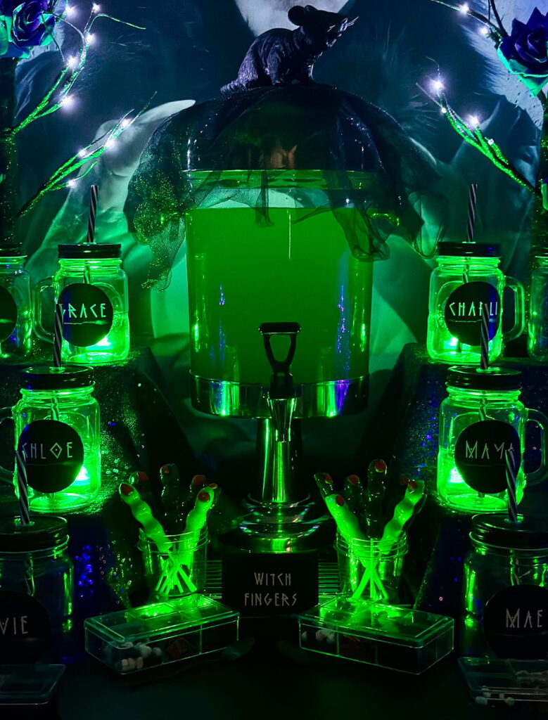 Witch-themed party drink table with glowing green jars and a drinks dispenser, set up for kids to serve themselves Witches' Brew at a spooky-cute witch party.