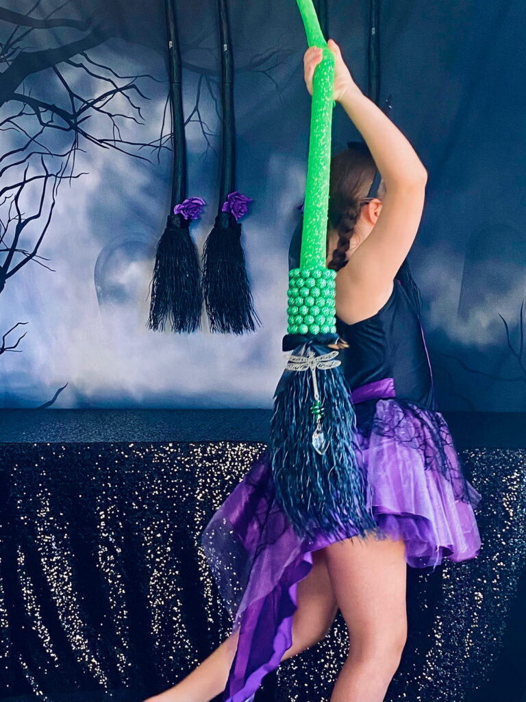 Young girl walking away holding a green decorated witches’ broom with beads and a crystal, part of a kids’ witch-themed party activity.