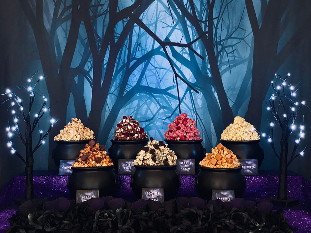 Witch-themed popcorn party table display with cauldrons piled high with different flavoured popcorn, a spooky tree backdrop, purple sequin tablecloth, and black led trees, perfect for a kids’ witch-themed party.