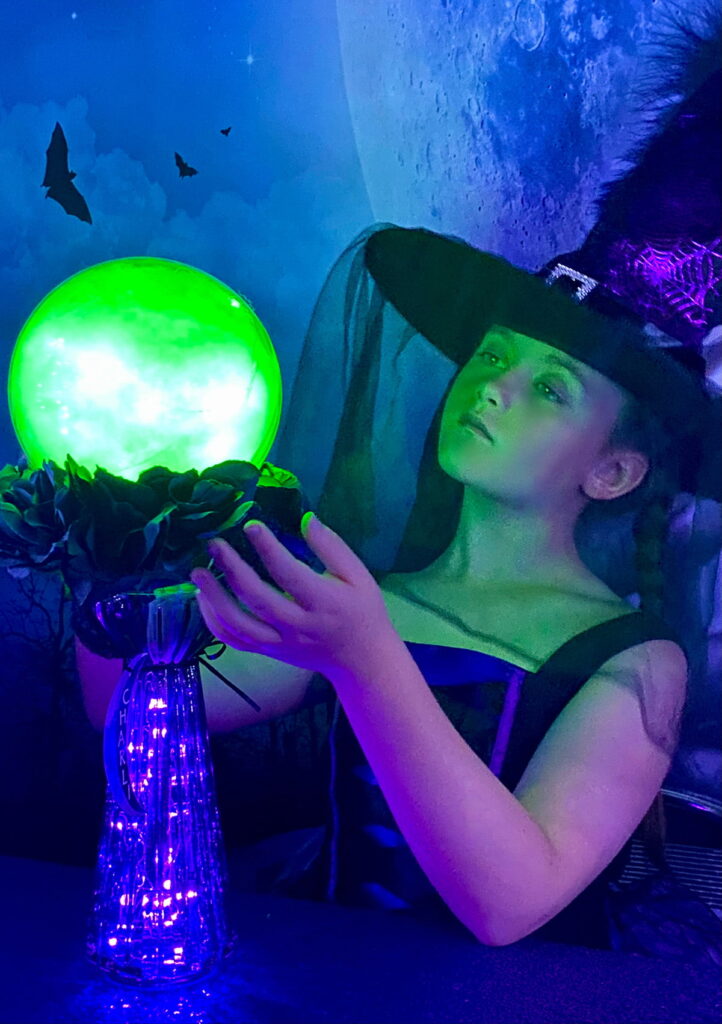 Young girl dressed as a witch admires her glowing crystal ball on a glass stand, looking up at it with delight during the kids’ crystal ball party.