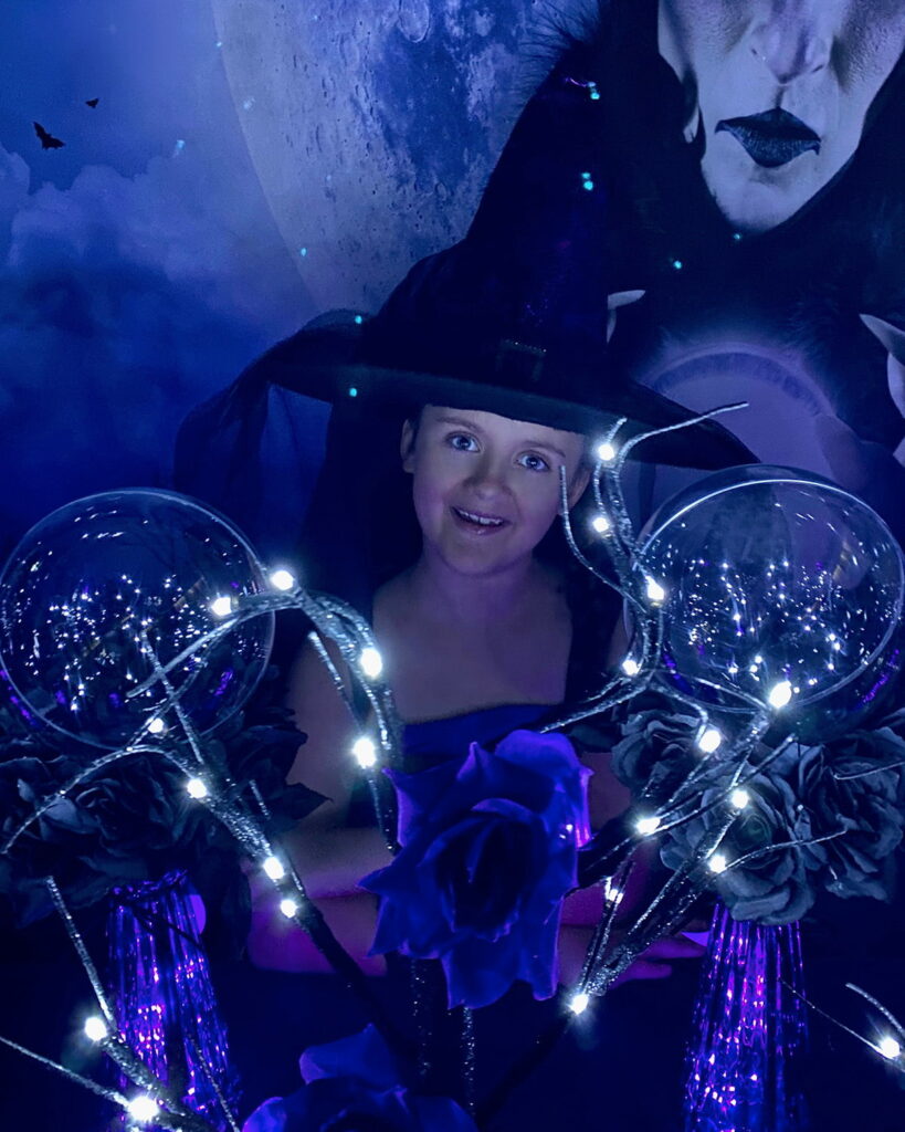 Young girl in a large witch hat sits at the crystal ball party table, smiling at the camera with crystal balls and decorations around her.