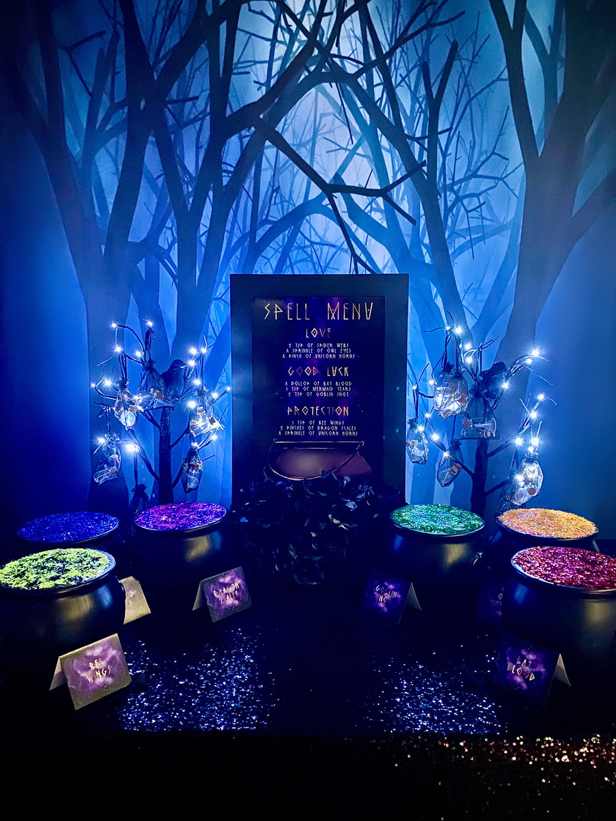 Overhead view of DIY spell jar party setup with black cauldrons filled with colourful glitter, glowing LED trees, and mini jars in black organza bags for kids’ craft activity.