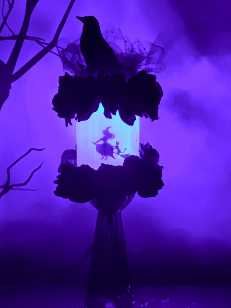 Glowing purple DIY Witch Lantern on a glass stand, featuring a witch on a broomstick, decorated with crow, tulle, and flowers on the lid.