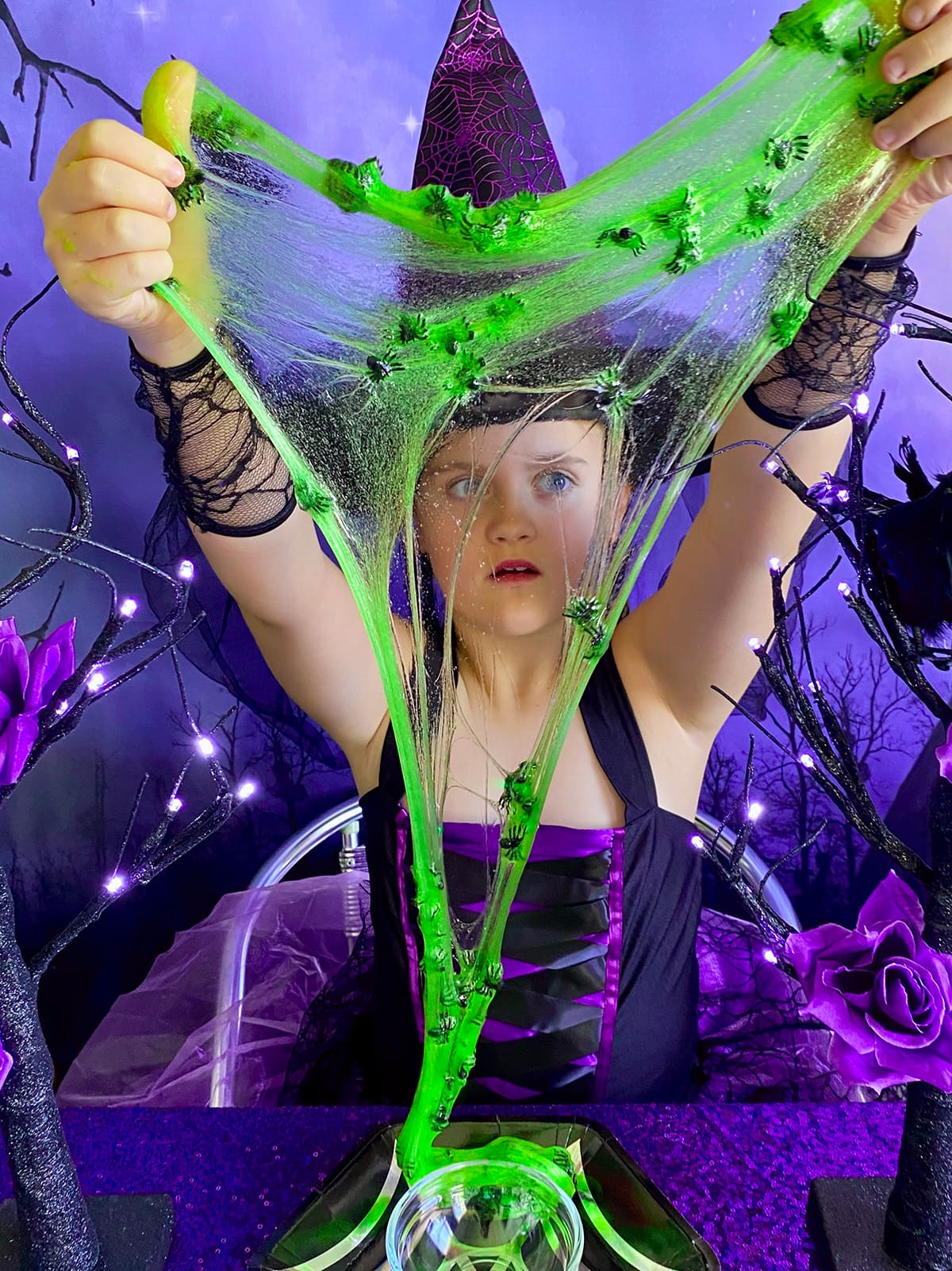 Young girl in a witch hat and dress looking spooked while stretching glowing green slime with black spiders at a cauldron slime party.