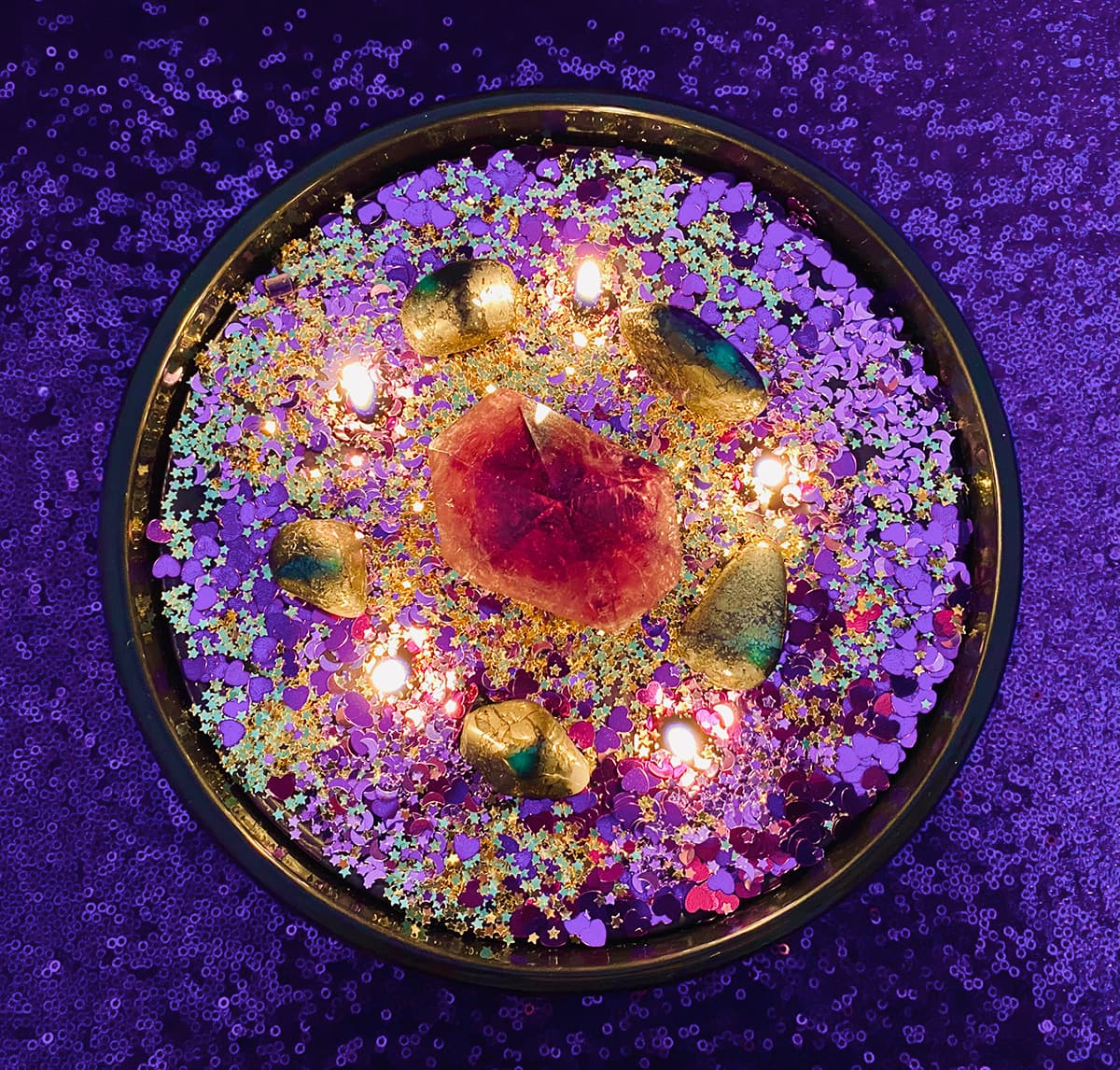 Overhead view of lit DIY crystal candle with purple point crystal, gold pyrite stones, and purple and gold glitter from kids’ candle decorating party.