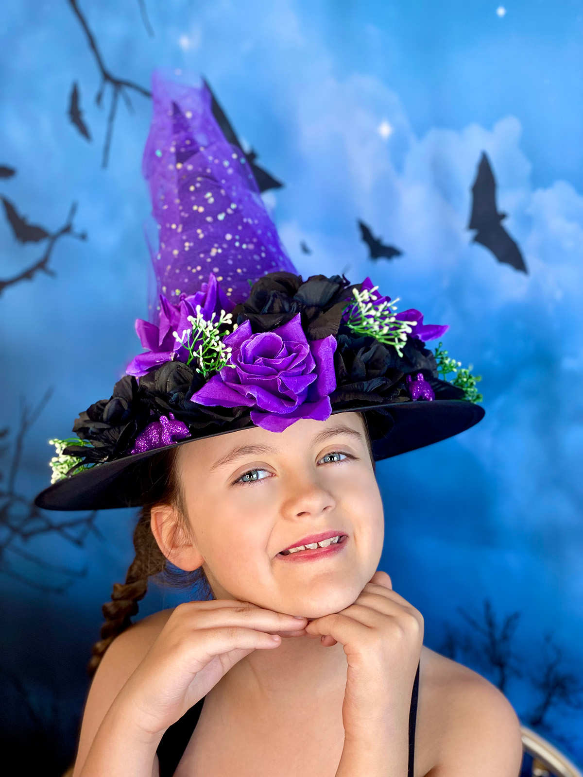 Young girl smiling with her hands under her chin, wearing a purple and black DIY witch hat decorated with glittery tulle, and black and purple roses.
