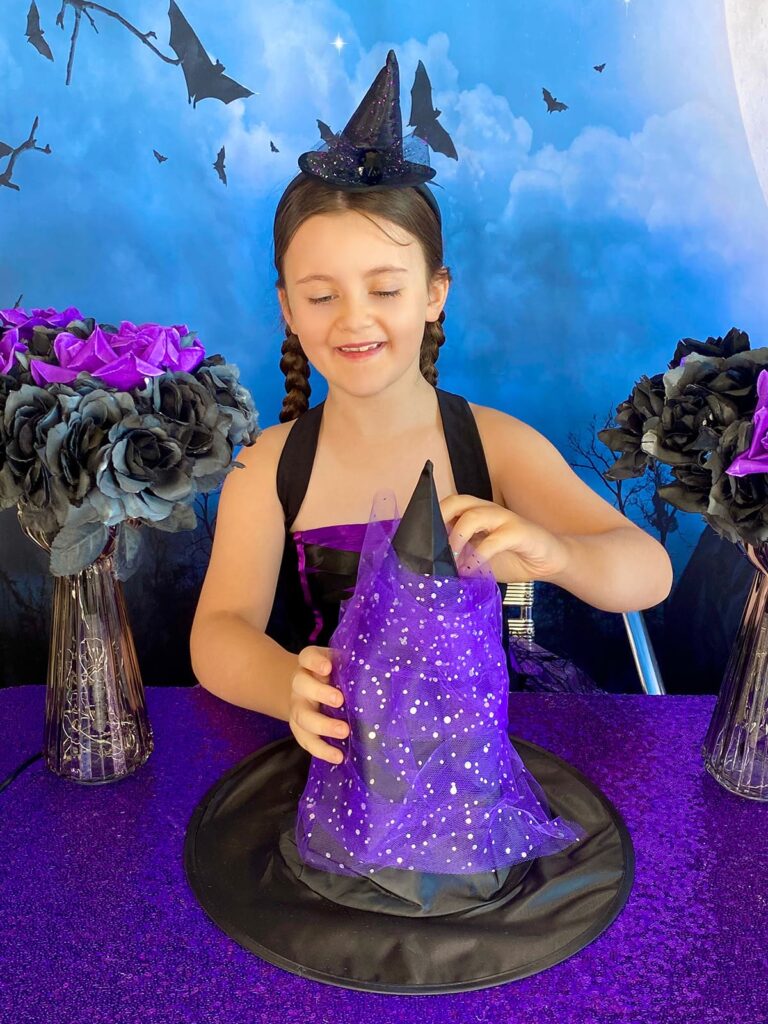 Young girl wearing a witch headband, smiling as she wraps purple tulle around a black DIY witch hat at a party table.