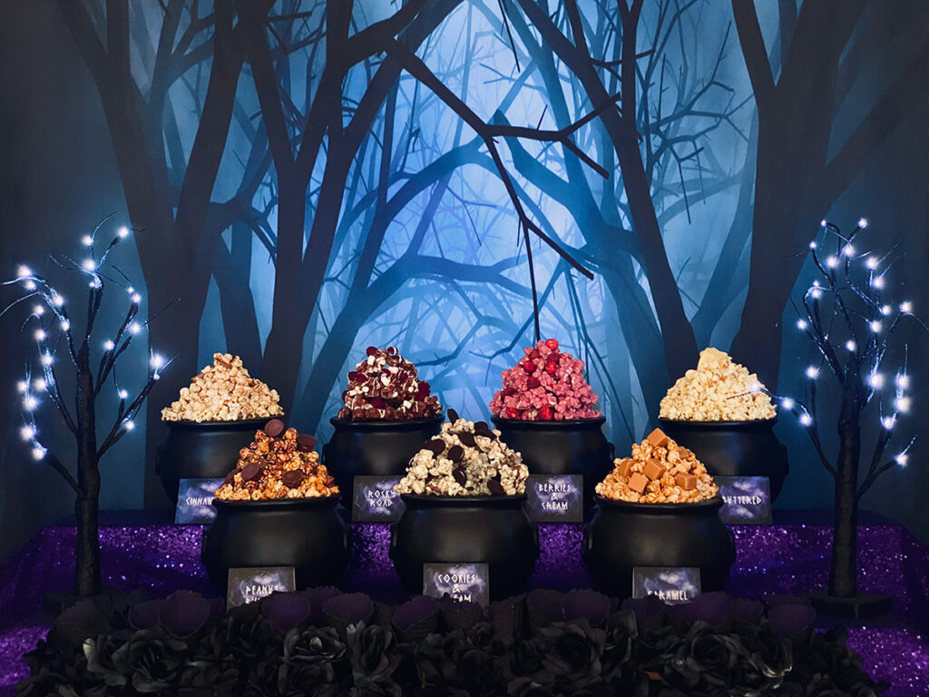Witch party popcorn table with seven cauldrons of popcorn, black waffle cones, purple sequin tablecloth, and spooky forest backdrop.