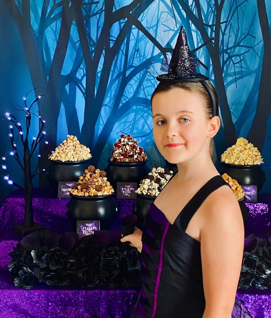 Young girl standing sideways in front of a witch party popcorn table, about to fill a waffle cone with popcorn, with a spooky forest backdrop visible behind her.