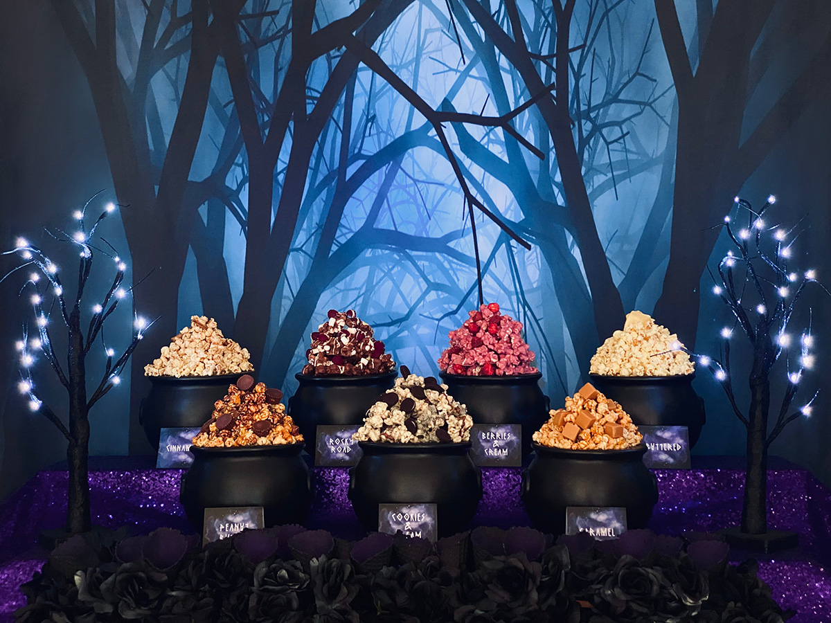 Witch party popcorn table with seven cauldrons of popcorn, black waffle cones, purple sequin tablecloth, and spooky forest backdrop.