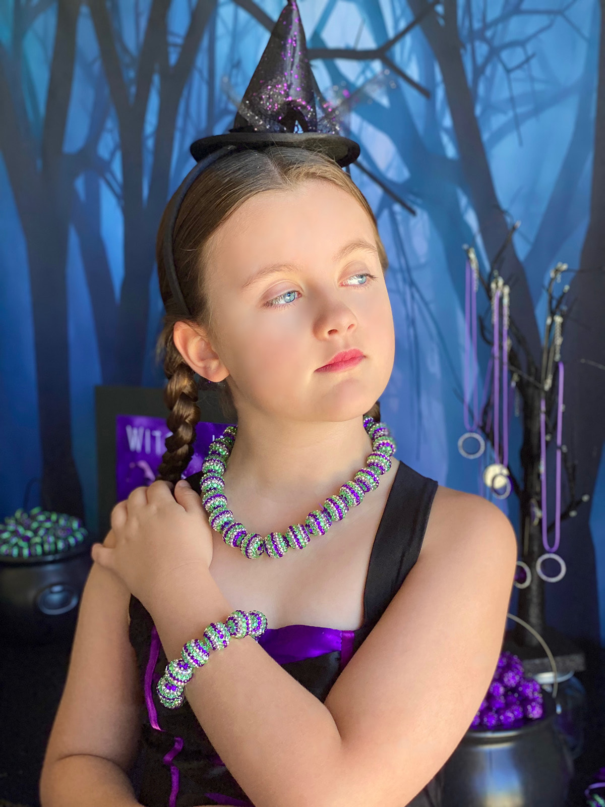 Young girl in a witch hat headband showing her DIY witch jewelry necklace and bracelet while looking to the side.