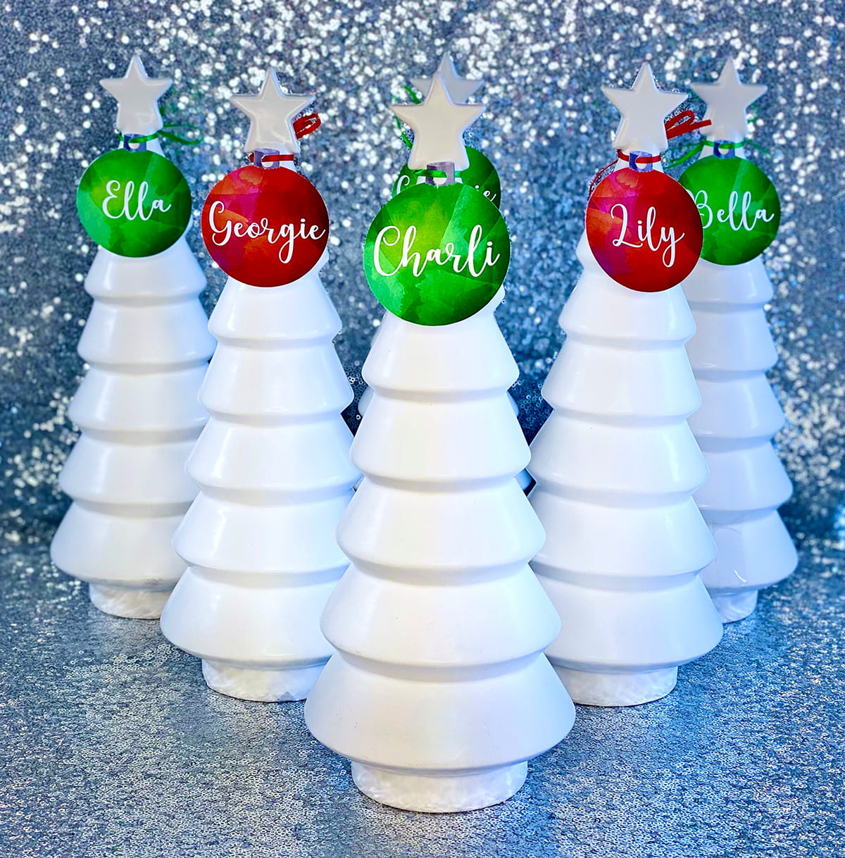 Five white ceramic Christmas trees with red and green personalised bauble name tags for a Crafty Christmas Tree kids’ party activity.
