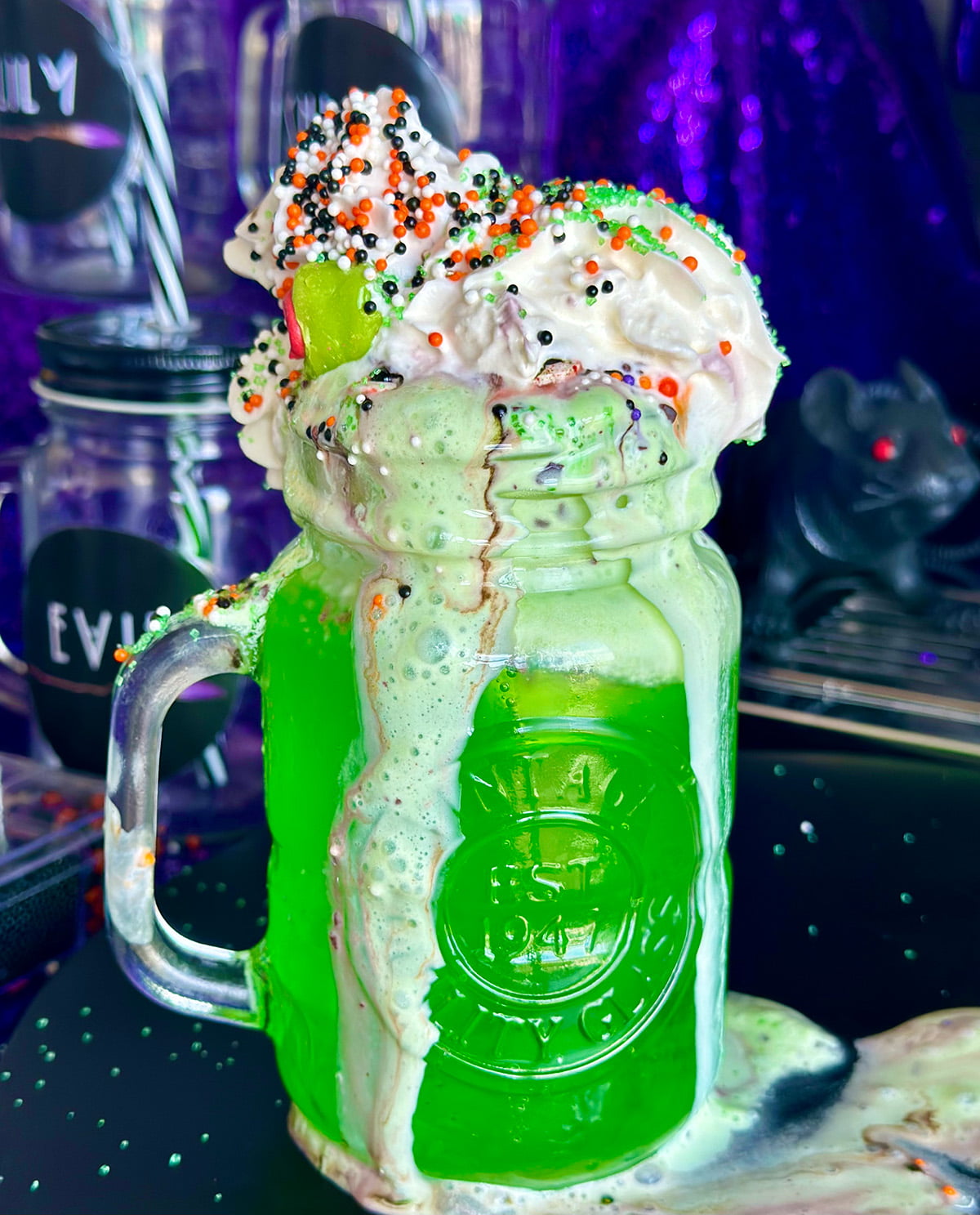 Creamy lime Spider Float in a mason jar with mint ice cream and whipped cream, topped with witch-themed sprinkles, overflowing onto the board underneath.