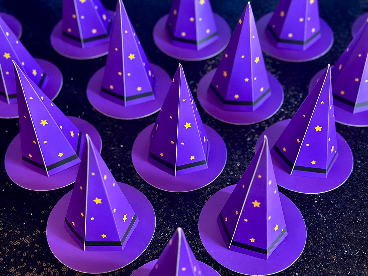 Mini purple cardboard witch hat party favour boxes decorated with yellow stars, displayed on a black sequin tablecloth for a kids’ witch-themed party.