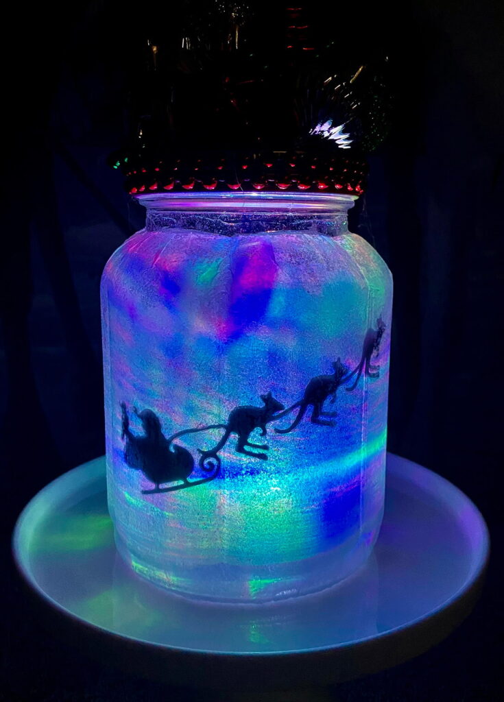 Close-up of a blue Christmas jar lantern with Santa’s sleigh led by flying kangaroos and glowing LED lights for a kids’ party activity.