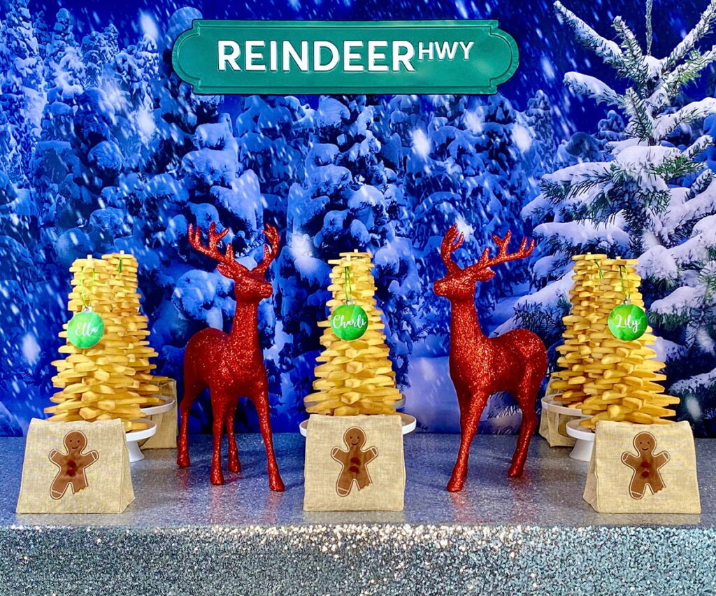 Christmas cookie tree party table with a snowy backdrop, silver sequin tablecloth, undecorated cookie trees, bauble name tags, and gingerbread man supply bags.
