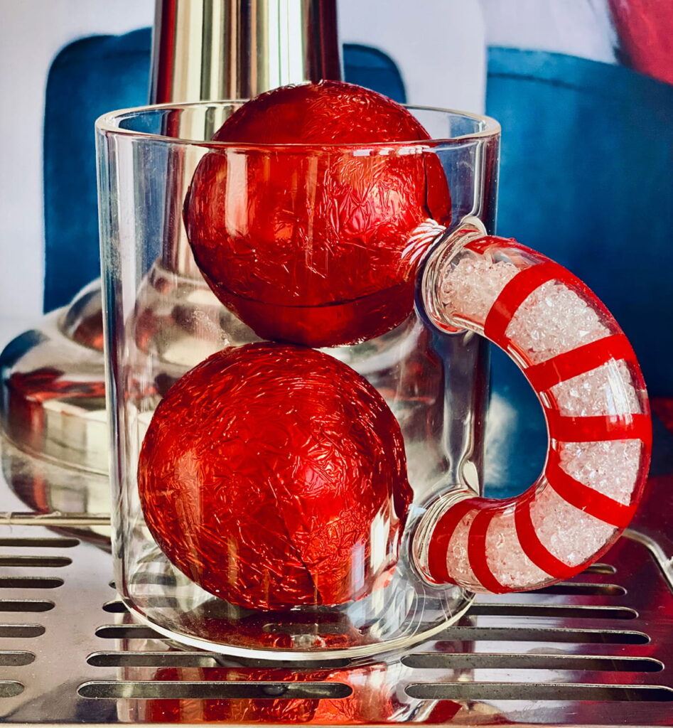 Close-up of a glass mug with a candy cane handle containing two red-foil chocolate hot cocoa bombs for a kids’ Christmas drink.
