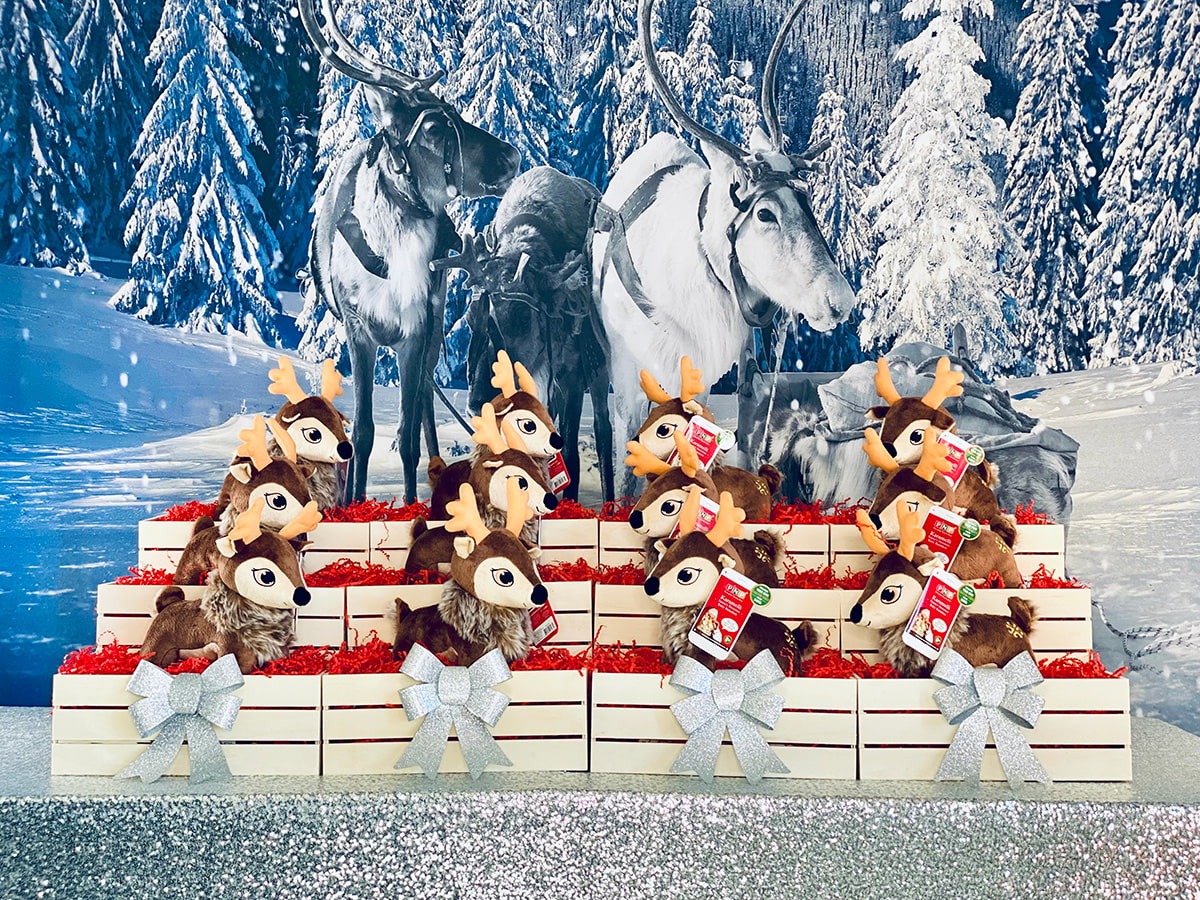 Adopt a Reindeer Christmas party table with plush reindeer in wooden crates filled with red paper shred, silver bows on the front crates, set on a silver sequin tablecloth with a snowy reindeer backdrop.