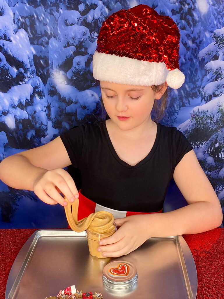 Young girl in red Santa hat making gingerbread-scented Christmas slime, twirling it in a jar.