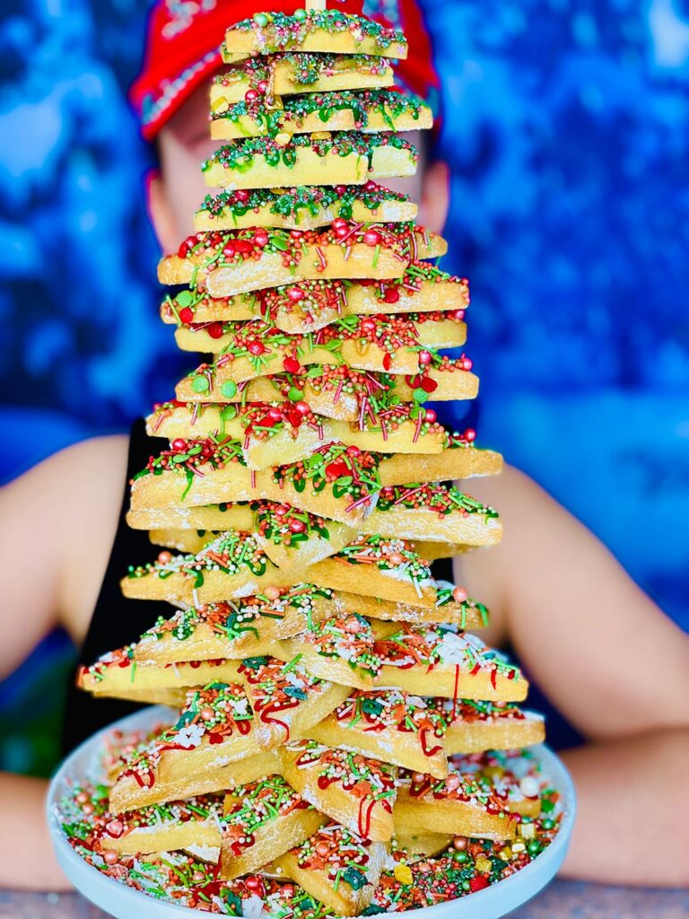 Close-up of a decorated Christmas cookie tree with festive sprinkles, with a young girl blurred in the background.