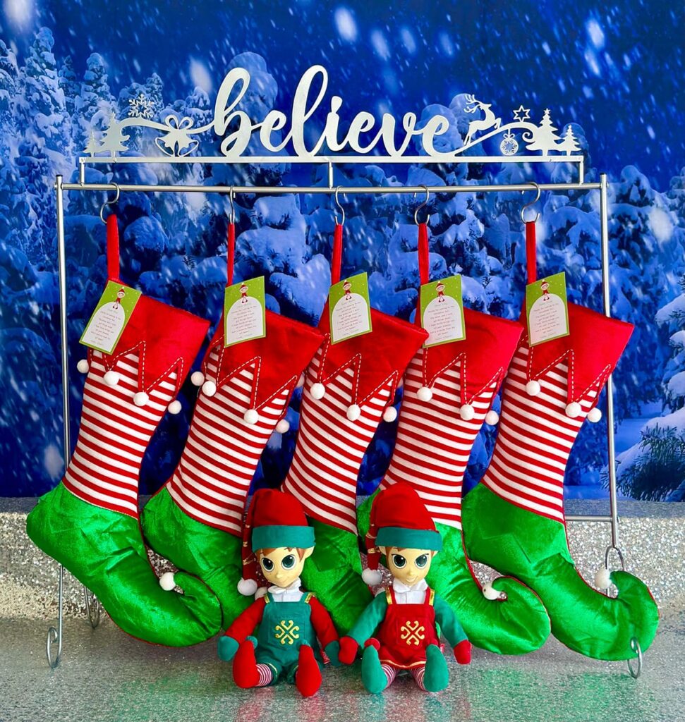 Elf-themed Christmas stockings hanging from a “Believe” stocking holder, each containing one portable North Pole elf as a kids’ party favour.