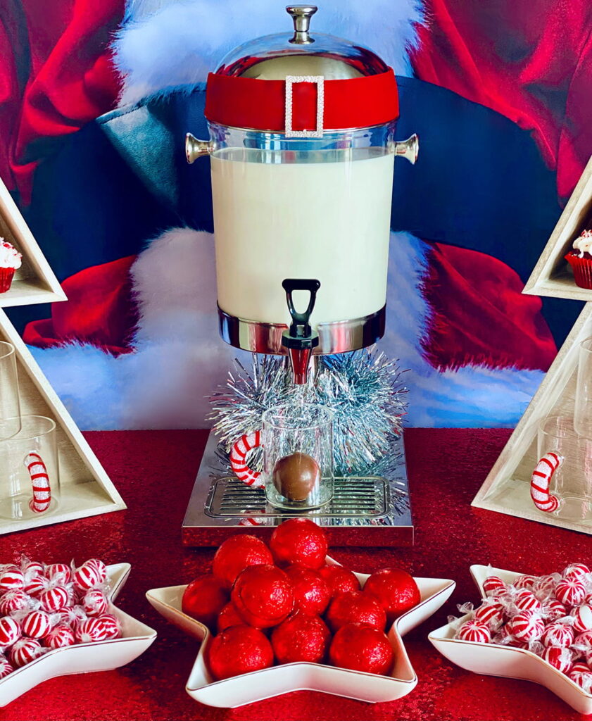 Kids’ Christmas hot cocoa setup with a milk dispenser, chocolate hot cocoa bombs in a glass, and festive holiday decorations.