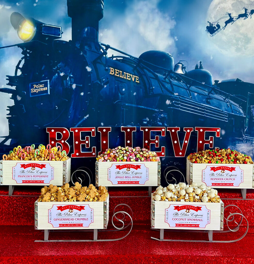 Kids’ Christmas party popcorn bar featuring a Polar Express train backdrop, red sequin-covered table, and sleigh-style crates holding assorted popcorn with train ticket labels.