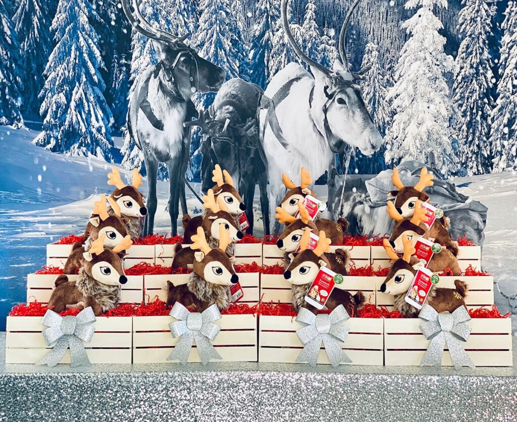 Kids' Christmas reindeer adoption party activity table with snowy backdrop, silver sequin tablecloth, and plush North Pole reindeer in crates.
