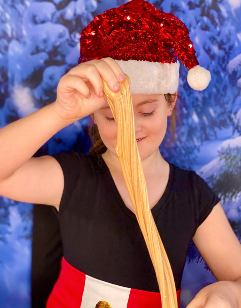 Young girl in red sequin Santa hat stretching gingerbread slime vertically and smiling while having fun at a kids’ Christmas slime party.