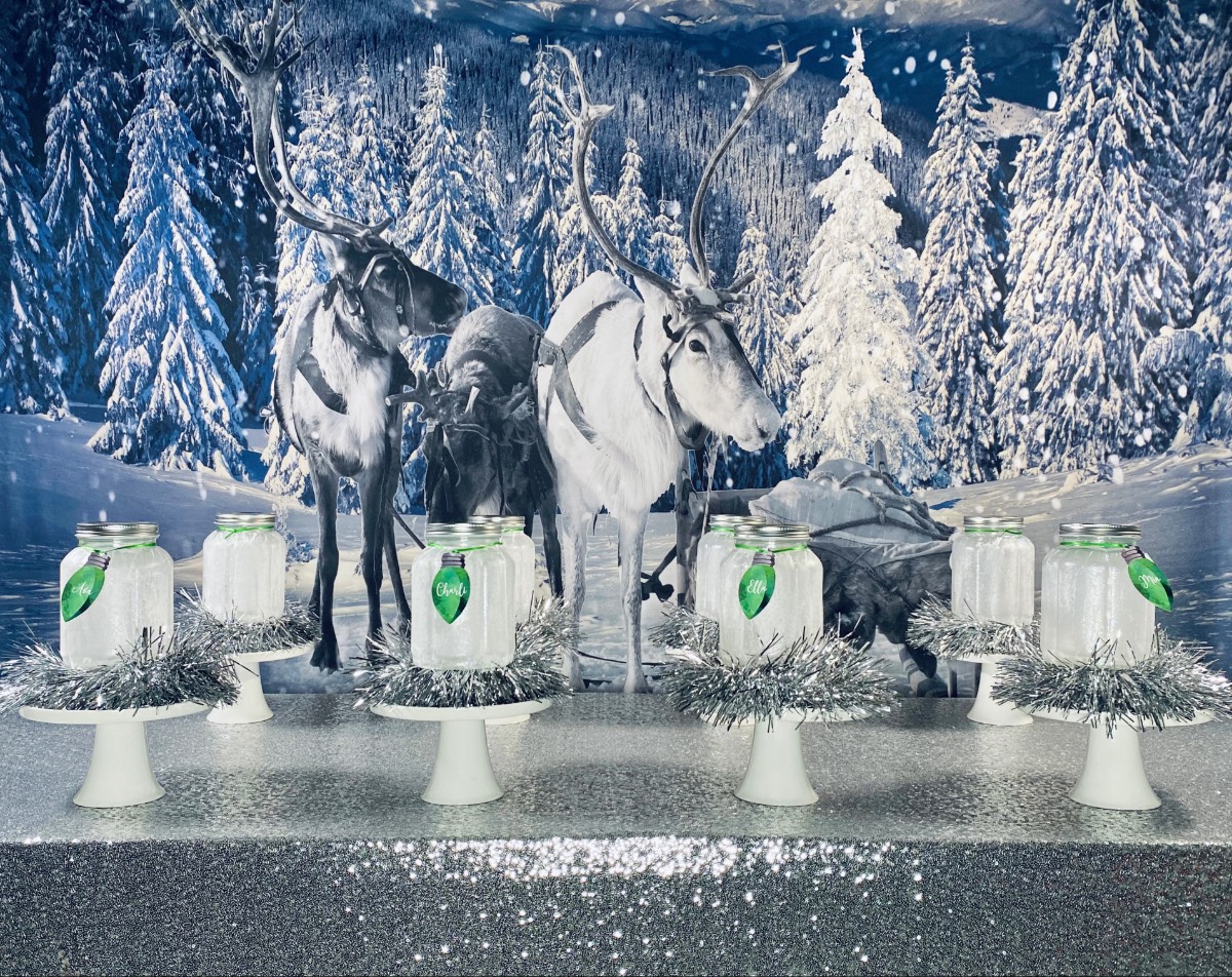 Christmas lantern craft party table with silver sequin tablecloth, snowy reindeer backdrop, mason jar lanterns, white cake stands, and green Christmas light name tags.