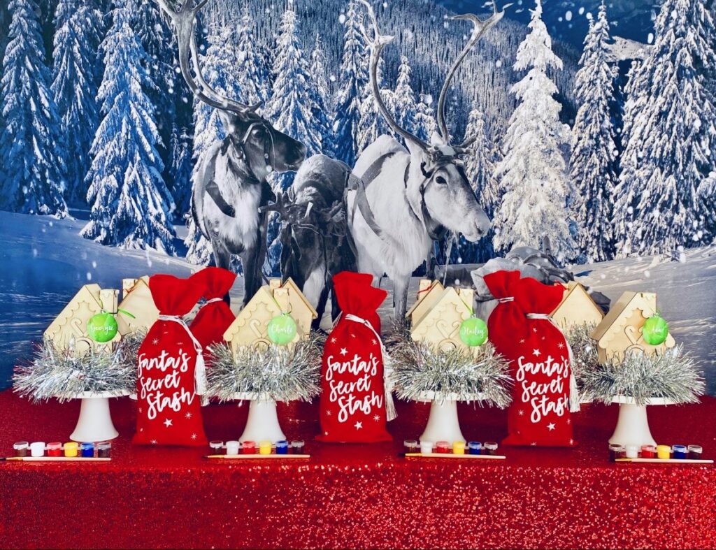 Christmas house decorating party table set up with red sequin tablecloth, snowy reindeer backdrop, undecorated wooden houses on cake stands with silver tinsel, red Santa’s Secret Stash supply bags, and paint pots.