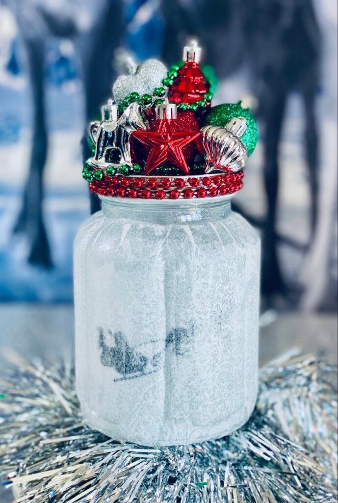Decorated Christmas fairy jar lantern with festive ornaments and a Santa sleigh silhouette inside a tissue-covered mason jar.