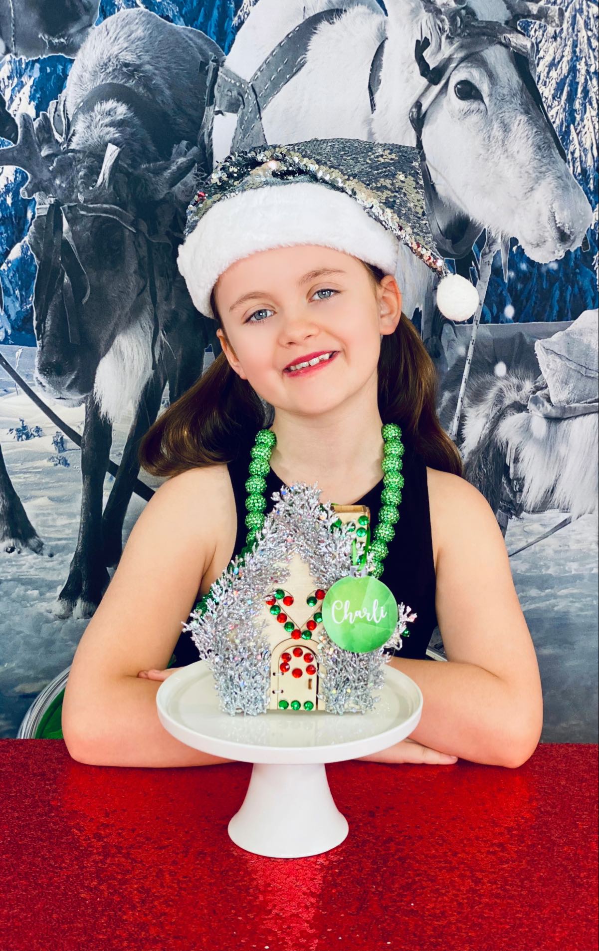 Smiling girl with folded arms sitting behind her decorated Christmas house on a cake stand, featuring silver foliage picks, red and green gems, and a personalised green bauble name tag.