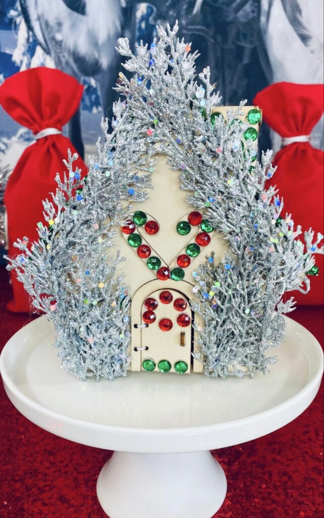 Decorated Christmas house on a cake stand with silver glittery picks and red and green gem trims, with red supply bags visible behind.